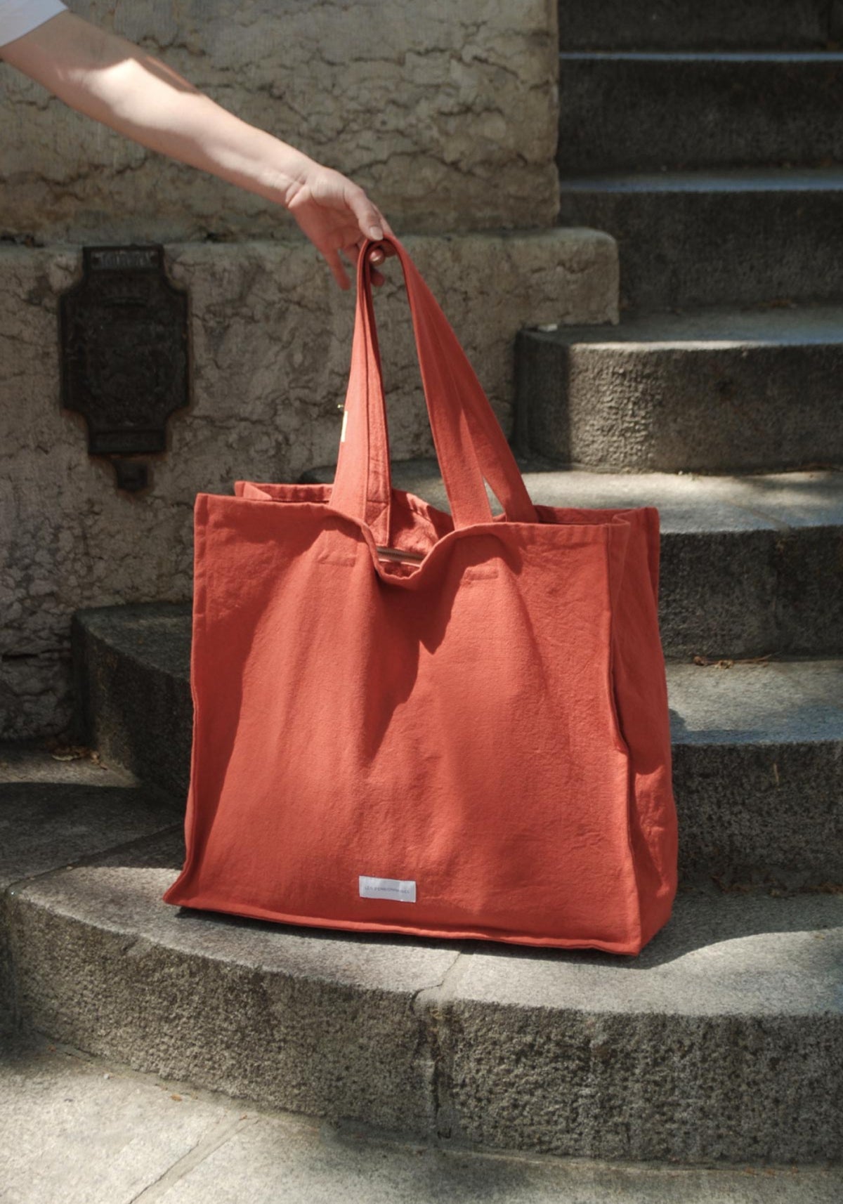 La femme tient le grand sac cabas orange flamboyant de chez Les Pensionnaires posé sur des escaliers