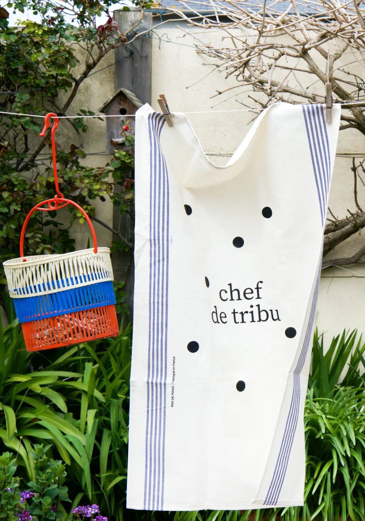 Le torchon en coton naturel Famille Chef de Tribu de chez Pied de Poule suspendu à un fil dehors avec un panier rempli d'épingles à linge